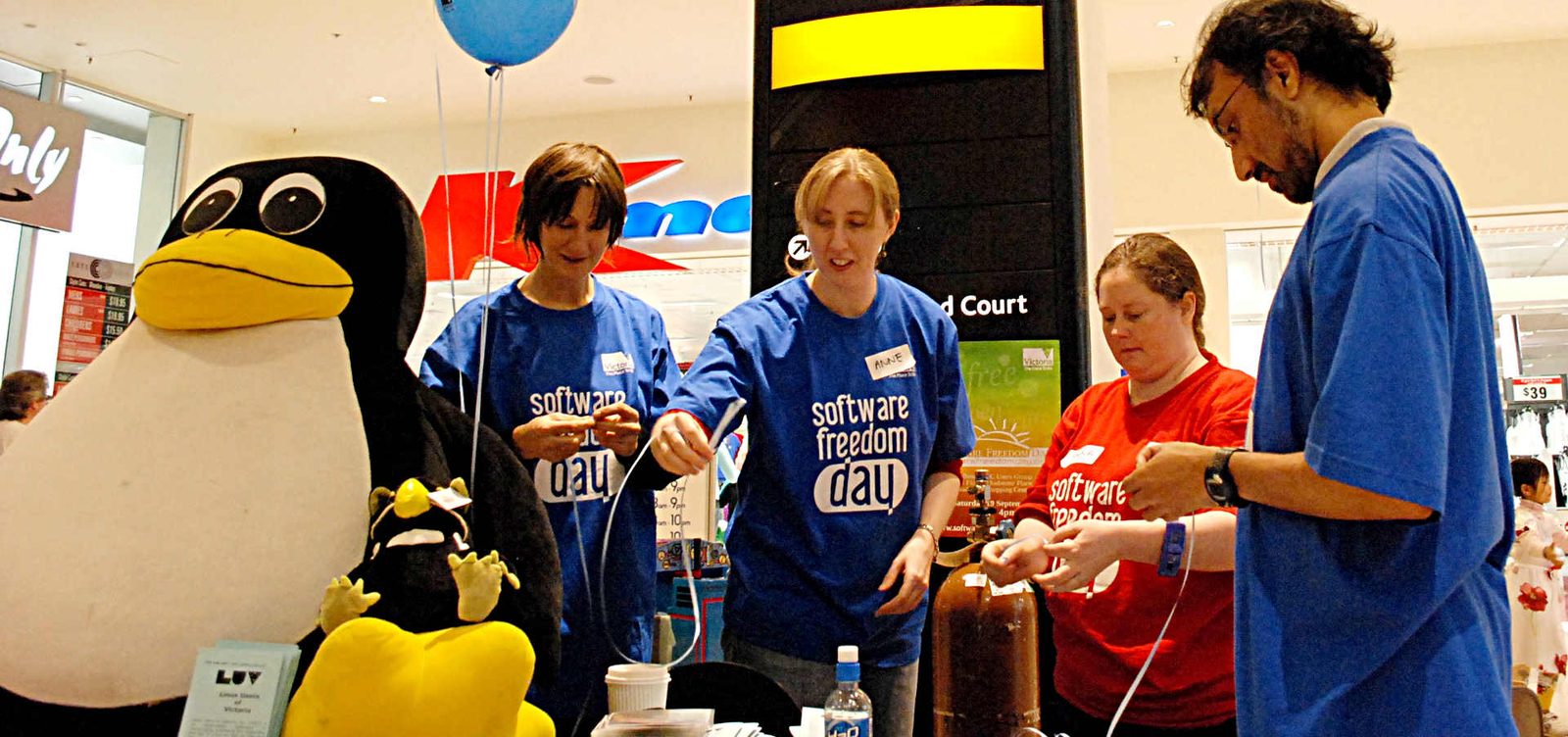 Volunteers in Software Freedom Day t-shirts running a stall in a shopping centre, with a large Tux penguin mascot beside them.